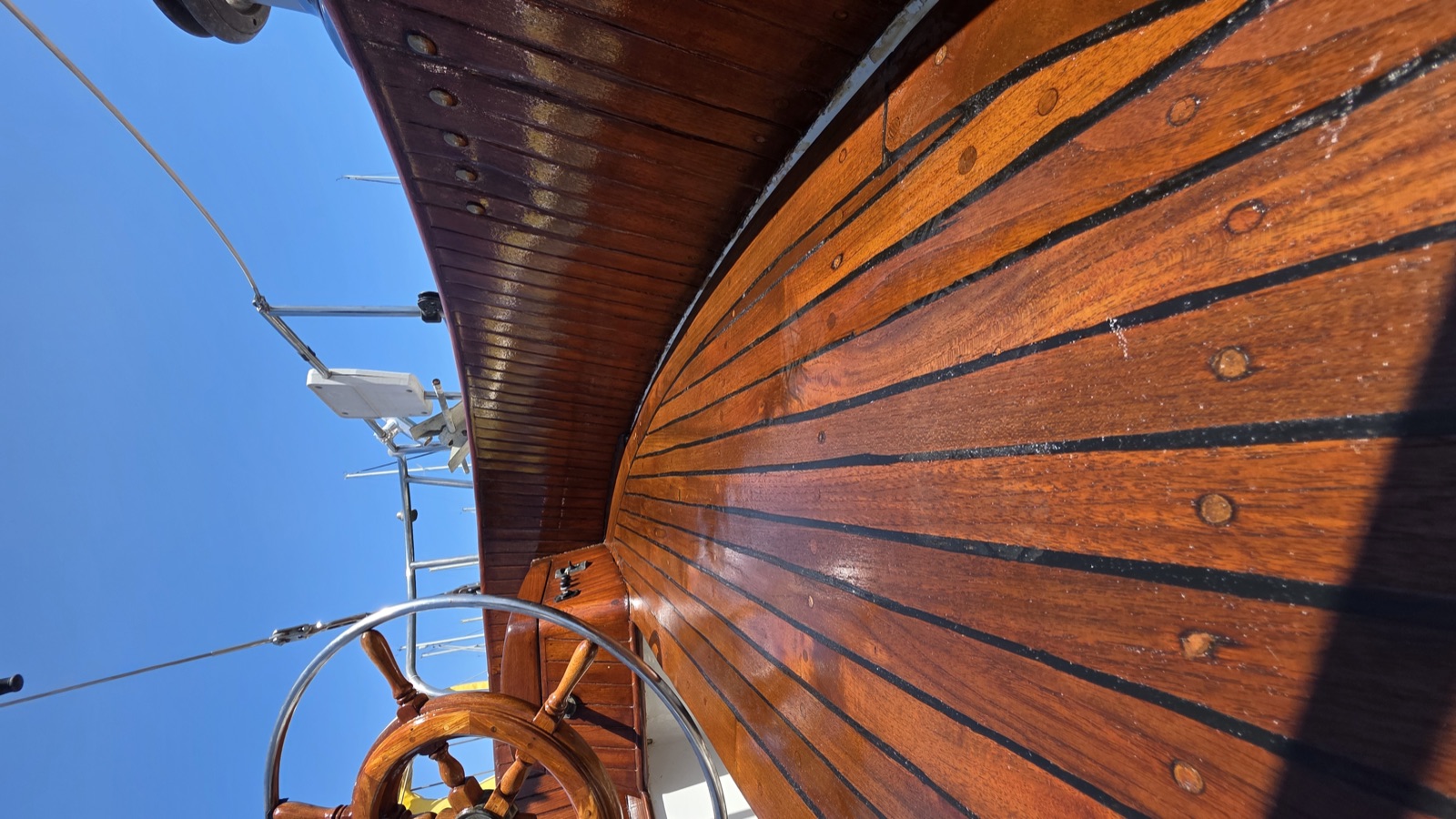 Looking up at glossy varnished teak rail and ship's wheel against blue sky